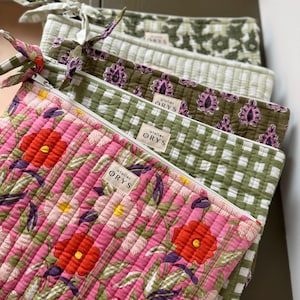 Può includere: Una pila di pochette in tessuto trapuntato con disegni floreali e fantasia. Le pochette hanno una chiusura a cerniera e un'etichetta con la scritta "atelier ORYS". I colori includono rosa, rosso, verde e bianco.