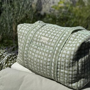 May include: A light green and white gingham patterned duffel bag. The bag has two handles and a small label that reads "ORYS". The bag is made of a quilted fabric. The bag is rectangular and appears to be filled with soft material.