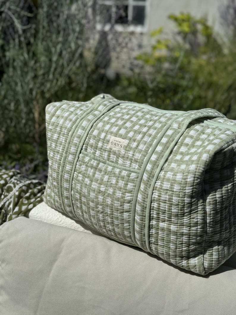 May include: A light green and white gingham patterned duffel bag. The bag has two handles and a small label that reads "ORYS". The bag is made of a quilted fabric. The bag is rectangular and appears to be filled with soft material.