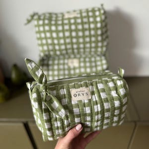 Può includere: Un beauty case a quadretti verdi e bianchi con chiusura a zip e un dettaglio in tessuto annodato. La borsa ha una forma rettangolare e una piccola etichetta con la scritta "atelier ORYS".