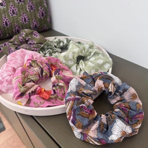 Könnte beinhalten: Eine Sammlung bunter Stoff-Haargummis in einer weißen Schale. Die Haargummis zeigen Blumen- und andere Muster in Rosa-, Grün- und Blautönen. Im Hintergrund ist eine passende gemusterte Tasche zu sehen.