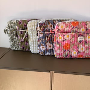 Puede incluir: Una fila de cinco fundas rectangulares acolchadas para portátiles con varios patrones, incluyendo estampados florales en rosa, azul y verde, así como un cuadro vichy verde claro. Cada funda tiene un cierre de cremallera y una etiqueta que dice atelier ORYS.