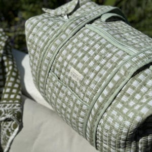 May include: A green and white checked travel bag with a zip closure and a handle. The bag is made of quilted fabric and has a small label with text. The bag is resting on a light-coloured surface.