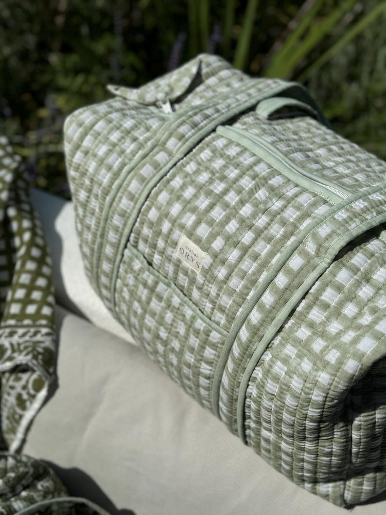 May include: A green and white checked travel bag with a zip closure and a handle. The bag is made of quilted fabric and has a small label with text. The bag is resting on a light-coloured surface.