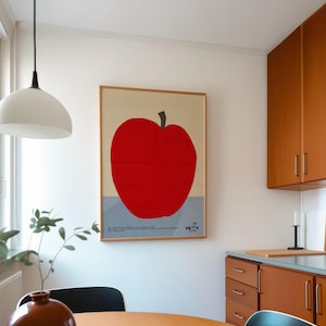 May include: A framed poster featuring a large red apple against a beige and light blue background. The poster is in a wooden frame and hangs on a white wall. The image is in a kitchen setting with a round wooden table and a brown vase.