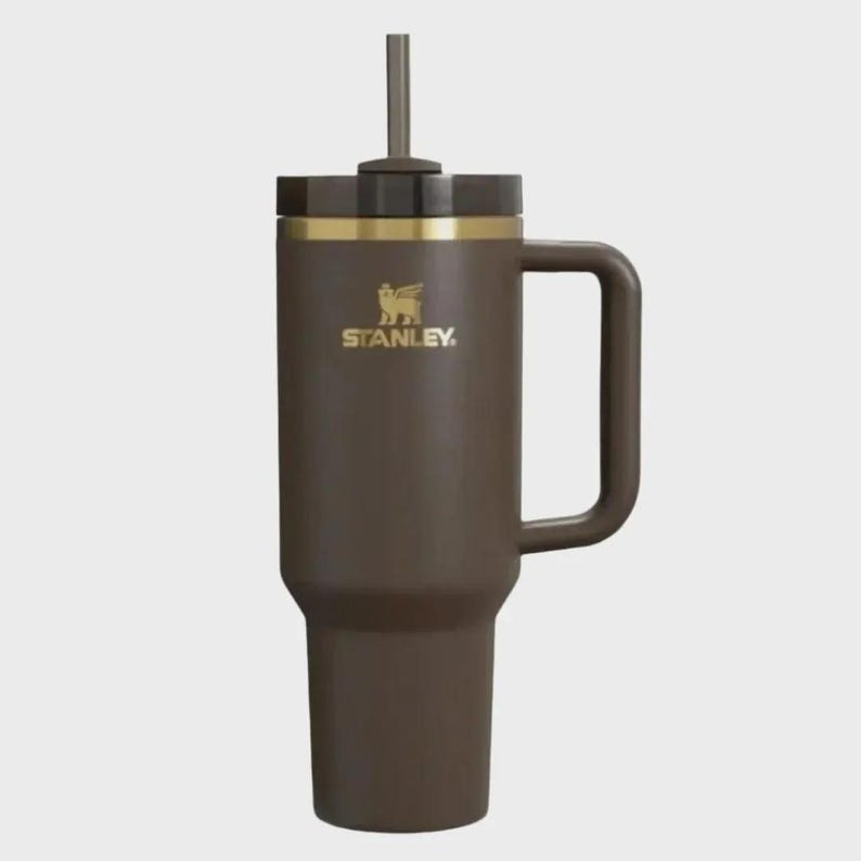 May include: A brown Stanley tumbler with a handle and straw. The tumbler has a gold band near the top and the Stanley logo in gold. The tumbler is designed to keep drinks cold or hot.