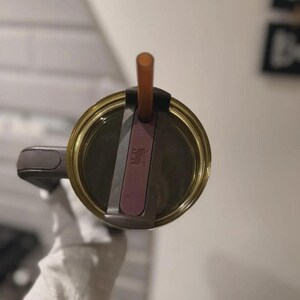 May include: A close-up view of a gold-colored tumbler with a brown straw. The lid is a dark gray color with a pink accent. The tumbler is held by a person wearing a white glove.