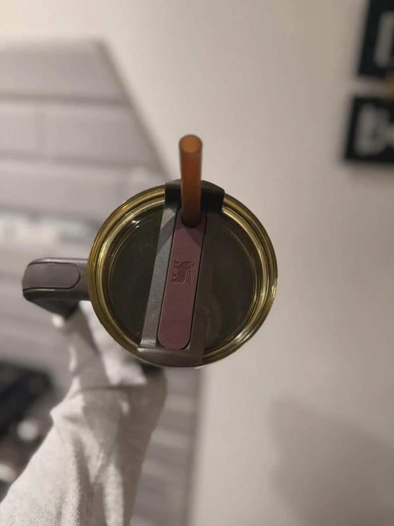 May include: A close-up view of a gold-colored tumbler with a brown straw. The lid is a dark gray color with a pink accent. The tumbler is held by a person wearing a white glove.