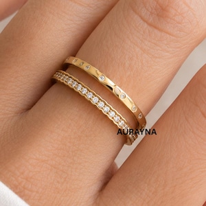 Puede incluir: Dos anillos dorados usados juntos en un dedo. El anillo superior presenta una serie de pequeñas piedras redondas espaciadas uniformemente en la banda. El anillo inferior es una banda más delgada incrustada con una fila continua de pequeñas piedras claras.