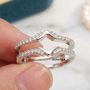 Puede incluir: Un protector de anillo plateado con un diseño en forma de V, incrustado con pequeñas piedras preciosas transparentes. El anillo está sostenido por una mano, con un fondo borroso.