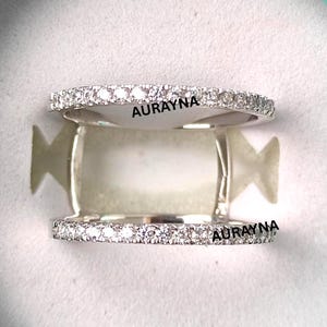 Könnte beinhalten: Zwei silberne Ringe mit einem Band aus kleinen, klaren Steinen. Die Ringe befinden sich in einer hellblauen Schachtel. Das Wort "AURAYNA" ist auf jedem Ring aufgedruckt.