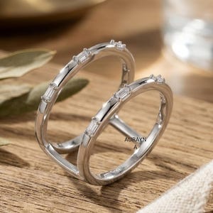 Può includere: Un anello a doppia fascia color argento con design a bypass, caratterizzato da piccole pietre trasparenti rettangolari taglio baguette incastonate sulla parte superiore di ogni fascia. L'interno riporta la scritta AURAYN A in carattere sans-serif.