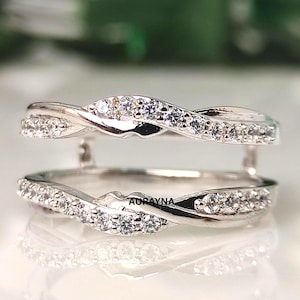 May include: A silver ring guard with a twisted design, encrusted with small, clear gemstones. The ring guard is designed to sit around a solitaire ring. The brand name "AURAYNA" is visible on the ring.