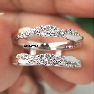 May include: A silver-toned ring guard featuring two curved bands with a twisted design, each set with small clear stones. The word AURAYNA is printed in black between the two bands. The metal has a polished finish and a reflective surface.