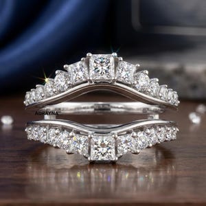 May include: A silver-toned ring guard set featuring two curved bands adorned with multiple round and princess-cut clear stones. The center of each band showcases a prominent princess-cut stone, with the text AURAYNA printed on the metal surface.