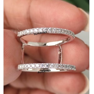 Könnte beinhalten: Nahaufnahme eines silbernen Rings mit zwei Bändern, die jeweils mit kleinen, klaren Edelsteinen besetzt sind. Die Bänder sind durch einen vertikalen Steg verbunden. Das Wort "AURAYNA" ist auf dem Ring zu sehen.