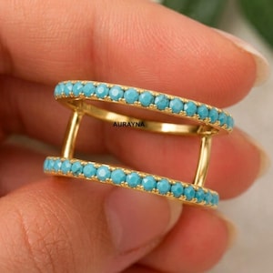 May include: A gold double-band ring featuring two parallel rows of small, round, bright turquoise stones. The bands are connected by two vertical gold bars, and the word AURAYNA is printed in small black letters on the inner gold bridge.