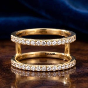 May include: A gold double-band ring featuring two parallel bands connected by vertical gold bars. Both bands are encrusted with small, round, clear pavé-set stones. The word AURAYNA is printed in small black letters on the inner band.