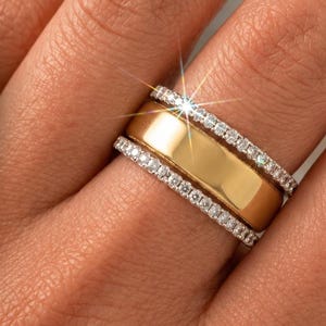 Könnte beinhalten: Ein goldfarbener Ringaufsatz kompatibel mit Oura Ring Gen 5, mit einem glatten Mittelband, das von zwei dünnen Reihen kleiner, klarer, funkelnder Steine flankiert wird. Der Text OURA RING und Compatible With Oura Ring Gen 5 ist links sichtbar.
