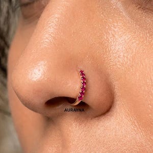 May include: A gold nose hoop featuring a row of five small, round, vibrant pink gemstones set along the outer curve. The jewelry is positioned in the nostril, and the word AURAYNA is printed in small black letters below the hoop.