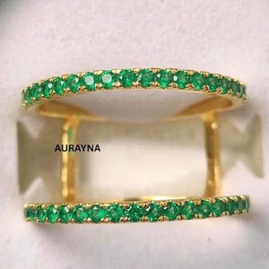May include: A double-band ring with a gold setting and a row of small, vibrant green gemstones on each band. The word "AURAYNA" is printed on the ring.