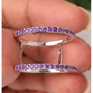 May include: A silver ring with two bands, each lined with small, round, purple gemstones. The ring is held up by two silver bars. The word "AURAYNA" is visible on the ring.