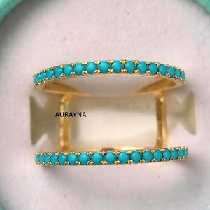 May include: A gold-colored double-band ring with a row of small, round turquoise stones set along the top and bottom bands. The ring has a modern, open design with the brand name "AURAYNA" visible.
