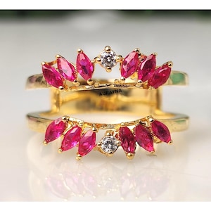 May include: A gold-colored ring with a double band design. The top band features marquise-cut, fuchsia-colored gemstones and a central clear gemstone. The bottom band mirrors the top. The text "AURAYNA" is visible.
