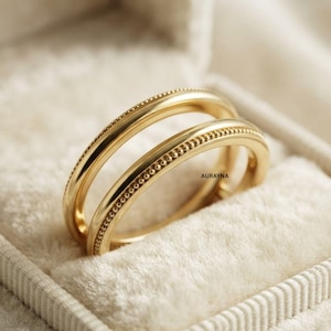 May include: A gold ring featuring a smooth, polished band paired with a second band detailed with a delicate beaded texture. The word AURAYNA is printed in small black letters on the inner surface of the beaded ring, showcasing a metallic finish.