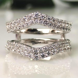 May include: Two silver-coloured, curved ring guards encrusted with numerous small, clear gemstones. The rings are stacked, with the text "AURAYNA" visible on the lower ring. The rings are set against a blurred, reflective background.
