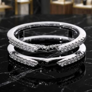 May include: A silver-toned ring guard featuring two parallel bands connected at the base. Each band is encrusted with small, clear, sparkling stones along the outer edges, meeting in a V-shaped point at the center. The letter A is visible.