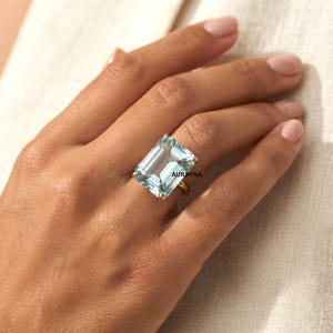 Könnte beinhalten: Ein Goldring mit einem großen, rechteckigen, hellblauen Edelstein im Smaragdschliff, der in einer Vier-Krappen-Fassung sitzt. Das Wort AURAYNA ist in kleinen schwarzen Buchstaben auf dem Stein gedruckt. Die Ringschiene ist dünn und aus poliertem Gold.