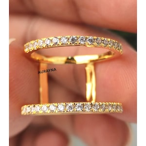 May include: A double-banded gold ring with a row of small, clear stones set along the top and bottom bands. The ring is held between fingers, with the brand name "AURAYNA" visible.