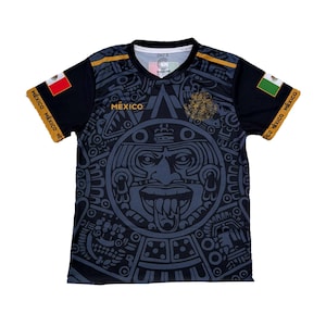 May include: Black and gray short-sleeve t-shirt featuring a large Aztec calendar design on the front. The shirt has the word "MEXICO" printed on the chest and the Mexican flag on the sleeves. Gold accents are present on the shoulders and sleeves.