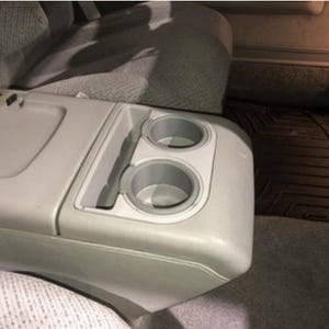 Pode incluir: Console de carro cinza claro com dois porta-copos e um compartimento de armazenamento. A consola faz parte de um interior de carro, com um assento cinza e um tapete preto no fundo.