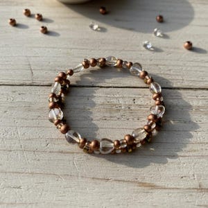 May include: A beaded bracelet with clear and copper-colored beads. The bracelet is made of clear, oval-shaped beads and small copper-colored beads. A small bowl with copper beads is in the background.