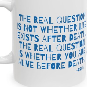 May include: White ceramic mug with a curved handle. The mug features blue text that reads: "THE REAL QUESTION IS NOT WHETHER LIFE EXISTS AFTER DEATH THE REAL QUESTION IS WHETHER YOU ARE ALIVE BEFORE DEATH -OSHA."