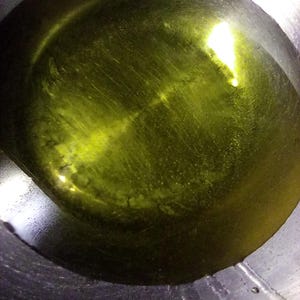 Op de afbeelding: Close-up van een roestvrijstalen pot gevuld met olijfolie. De olie heeft een levendige geelgroene tint, die licht reflecteert over het oppervlak. De binnenkant van de pot vertoont subtiele cirkelvormige patronen.
