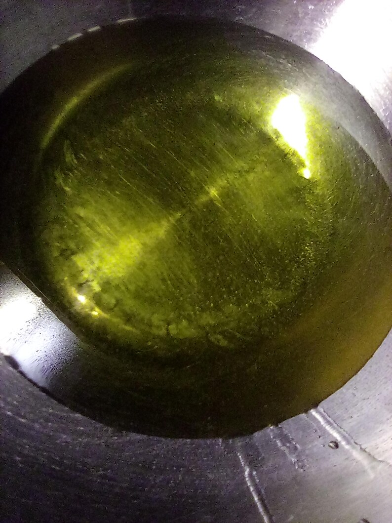 Op de afbeelding: Close-up van een roestvrijstalen pot gevuld met olijfolie. De olie heeft een levendige geelgroene tint, die licht reflecteert over het oppervlak. De binnenkant van de pot vertoont subtiele cirkelvormige patronen.