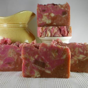 May include: Handmade soap bars with a layered design. The soap features a blend of pink, brown, and cream colors, creating a marbled effect. The bars are arranged on a white surface, with a yellow pitcher in the background.