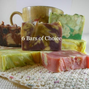 May include: Six bars of handmade soap in various colors and patterns, including brown, green, yellow, purple, and pink. The soap bars are stacked on top of each other and are sitting on a crocheted doily. The text "6 Bars of Choice" is visible in the center of the image.