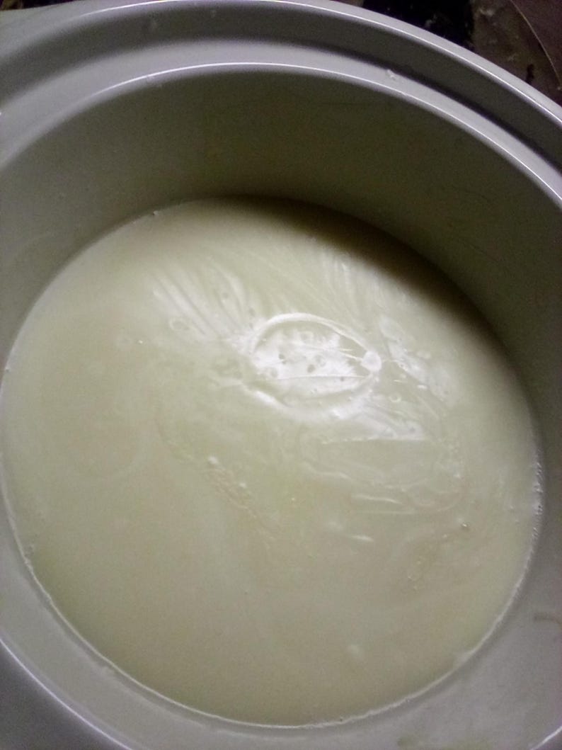 Op de afbeelding: Een close-up van een lichtgekleurde vloeistof in een grijze keramische slowcooker. De vloeistof heeft een romige, ondoorzichtige uitstraling met subtiele oppervlaktestructuren en een paar bellen. De slowcooker is rond met een brede rand.