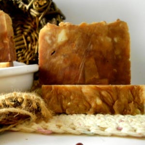May include: Two rectangular bars of amber-colored soap with visible textured inclusions and a marbled appearance. The soap bars are stacked, with one standing upright and the other lying flat, showcasing a rustic, uneven surface and earthy tones.