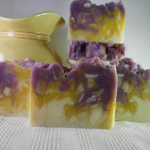 May include: Artisan soap bars with a layered design. The soaps feature a blend of colors, including purple, yellow, and white. The bars have a textured top and a smooth, creamy base. A yellow pitcher is in the background.