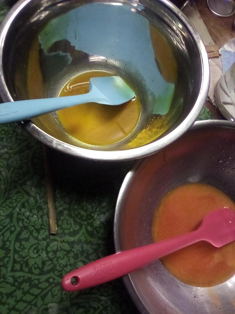 Op de afbeelding: Twee roestvrijstalen mengkommen met kleurrijke inhoud. E&eacute;n kom bevat een gele vloeistof met een lichtblauwe spatel, de andere een oranje vloeistof met een roze spatel. De kommen staan op een groen oppervlak met een patroon.