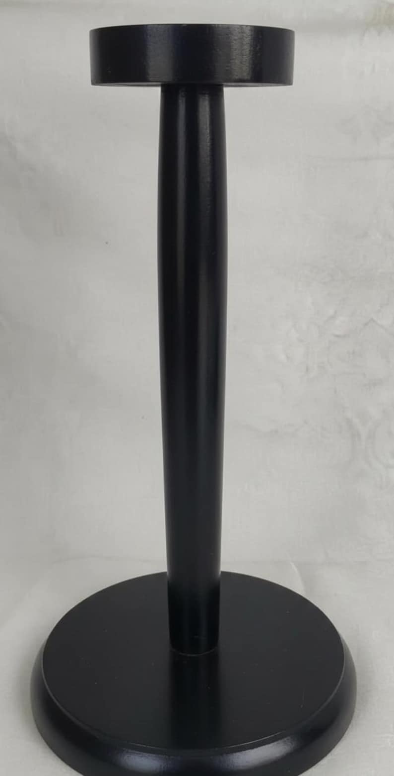 May include: A black wooden display stand featuring a circular base, a central vertical post, and a flat circular top platform. The stand has a smooth, matte finish and is designed to hold items securely in an upright position for display.
