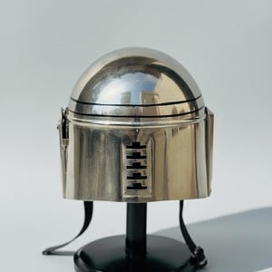 May include: A silver-colored helmet with a rounded top and a cylindrical base. The helmet has black horizontal lines and a series of rectangular cutouts. It is displayed on a black stand with a circular base.