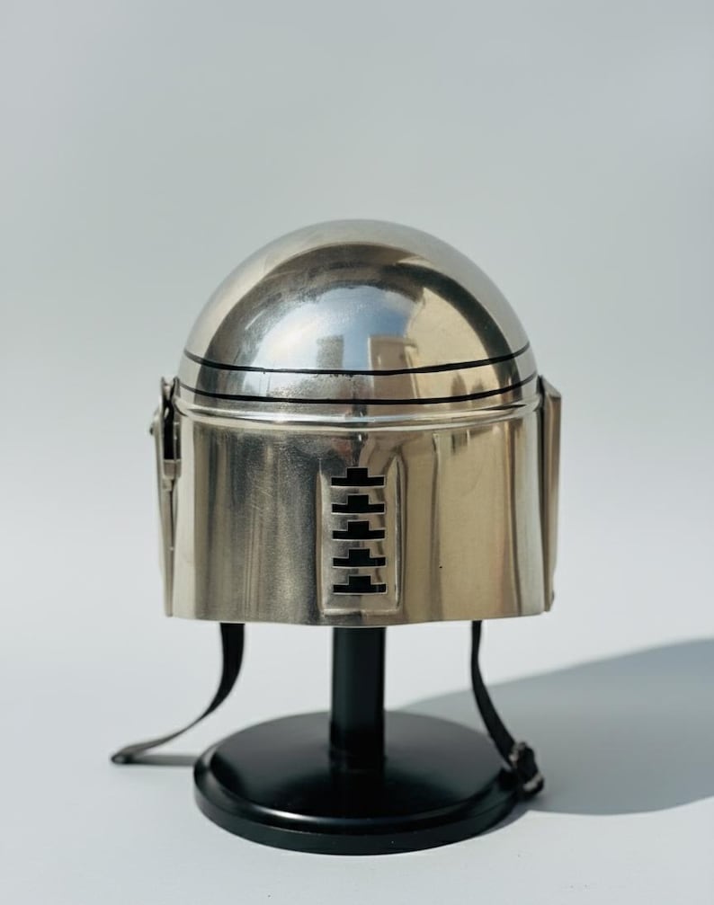 May include: A silver-colored helmet with a rounded top and a cylindrical base. The helmet has black horizontal lines and a series of rectangular cutouts. It is displayed on a black stand with a circular base.