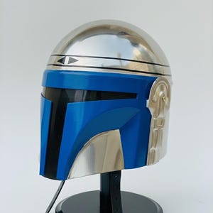May include: A metallic silver and blue helmet, likely for cosplay or display. The helmet features a rounded top, a blue visor area, and silver accents. It is mounted on a black stand with a strap.