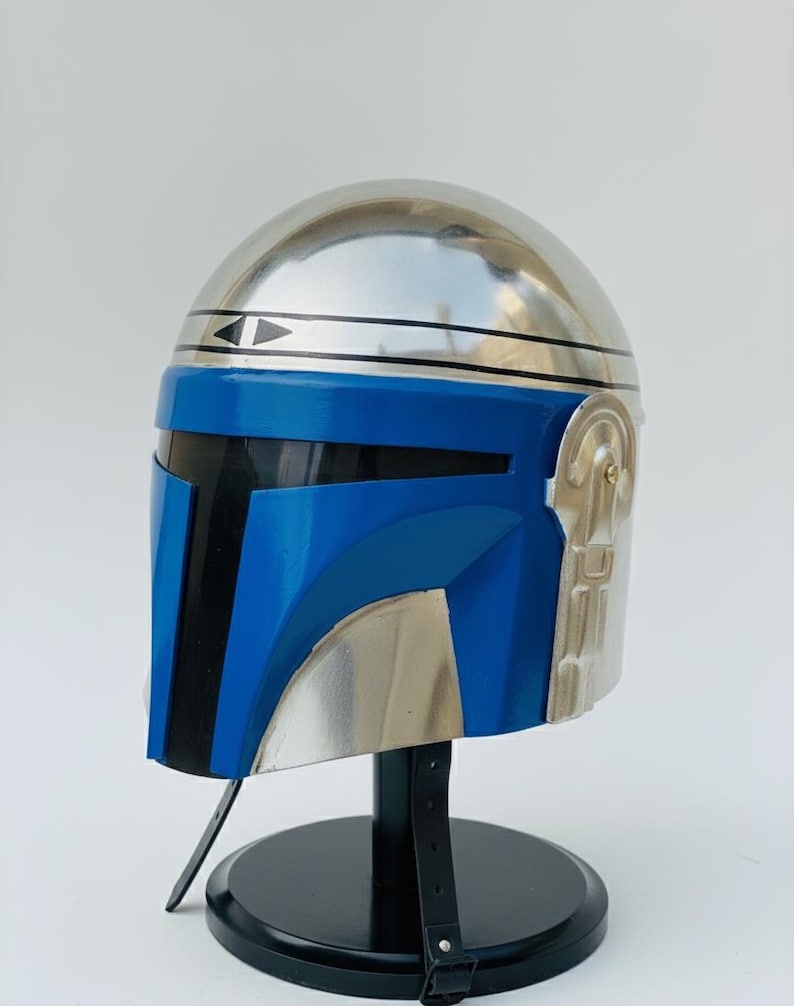 May include: A metallic silver and blue helmet, likely for cosplay or display. The helmet features a rounded top, a blue visor area, and silver accents. It is mounted on a black stand with a strap.